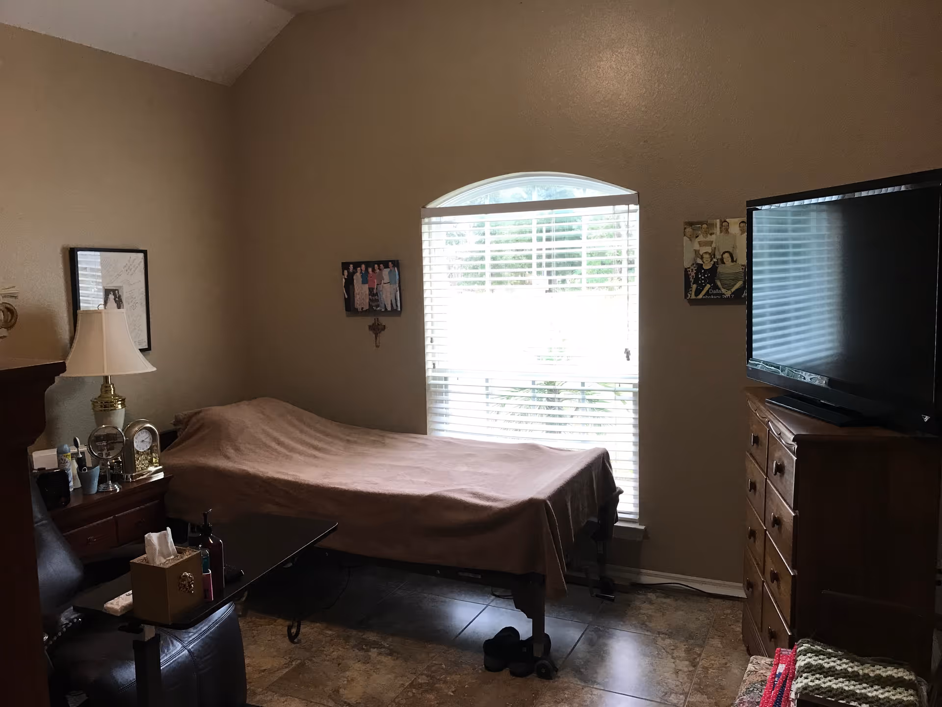 A small bedroom with a single bed covered by a brown blanket positioned against a wall with a window that has white blinds. There is a wooden dresser with a large flat-screen TV on top to the right of the bed. On the left side, there is a nightstand with a lamp, clock, and other small items. The floor is tiled, and there are a few framed pictures on the walls.