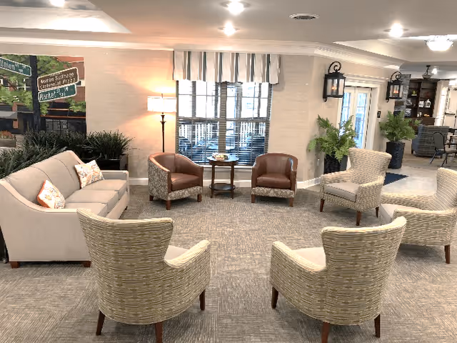 Well-lit senior living lounge with a sofa and multiple armchairs arranged in a circle, side tables, plants, and a window with a striped valance.