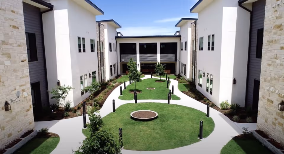 Outdoor courtyard area of a senior living facility with green grass, small trees, and a circular planter in the center. The courtyard is surrounded by two-story buildings with white and stone exterior walls and multiple windows. A clear blue sky is visible above.