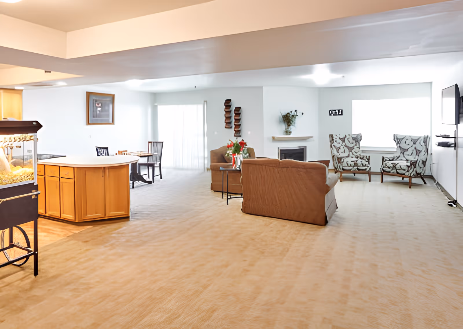Bright open communal living area with sofas and armchairs arranged around a fireplace and TV, adjacent to a small kitchen island.
