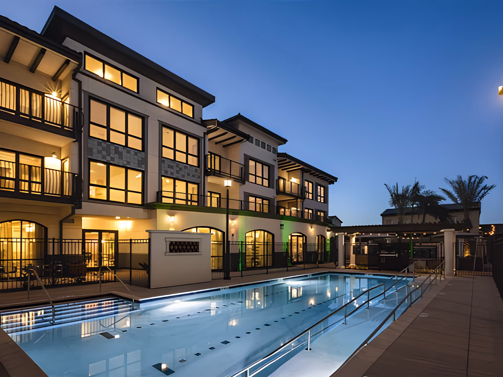 Exterior view of a three-story building at dusk with lit windows and a swimming pool in the foreground.