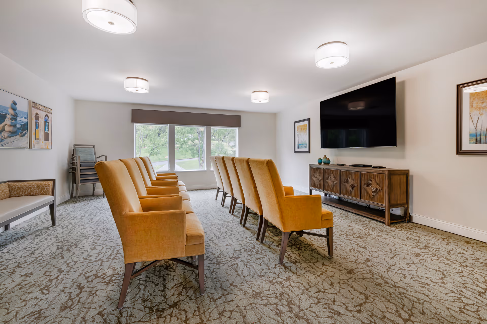 A bright room with two rows of mustard yellow upholstered chairs facing a large flat-screen TV mounted on the wall. The room has patterned carpet, white walls with framed artwork, and large windows letting in natural light. There is a wooden cabinet under the TV and a bench along the left wall.