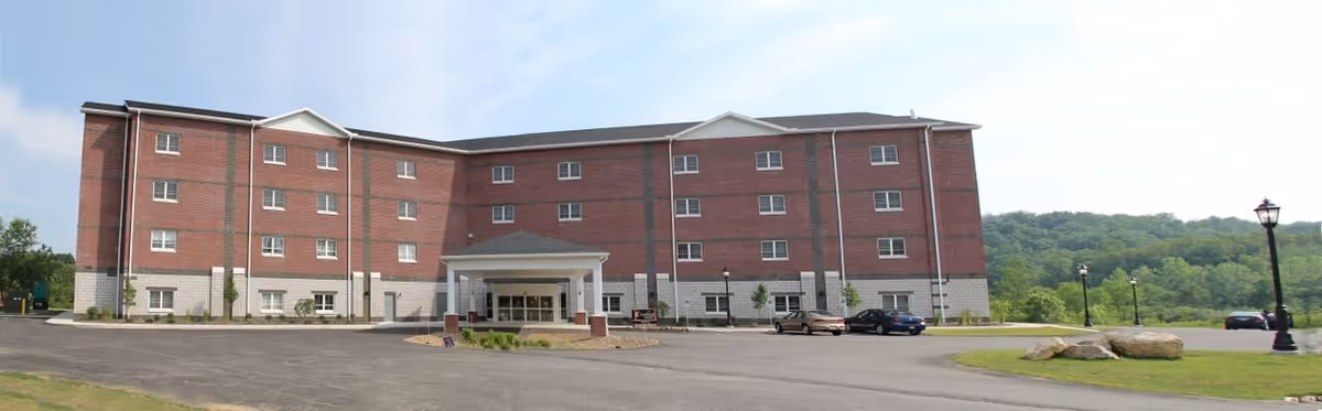 Exterior view of a four-story brick senior living facility building with a covered entrance, surrounded by a parking lot and green landscape with trees and hills in the background under a clear sky.