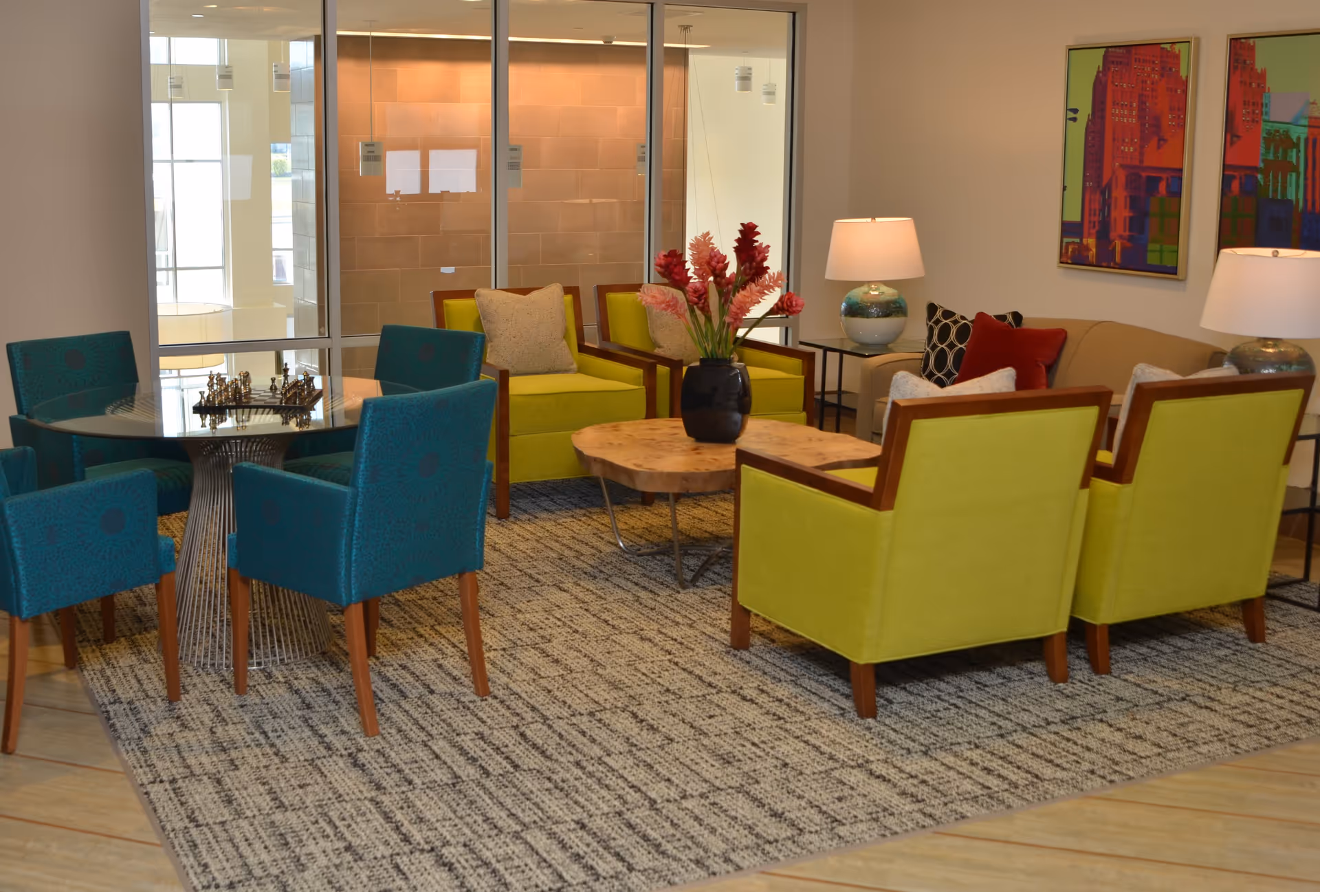 A cozy seating area in a senior living facility featuring a beige sofa with decorative pillows, four lime green armchairs, and a round glass table with four teal chairs. A wooden coffee table with a black vase holding red flowers is centered on a patterned rug. Two table lamps with blue and white bases and colorful abstract artwork hang on the wall.