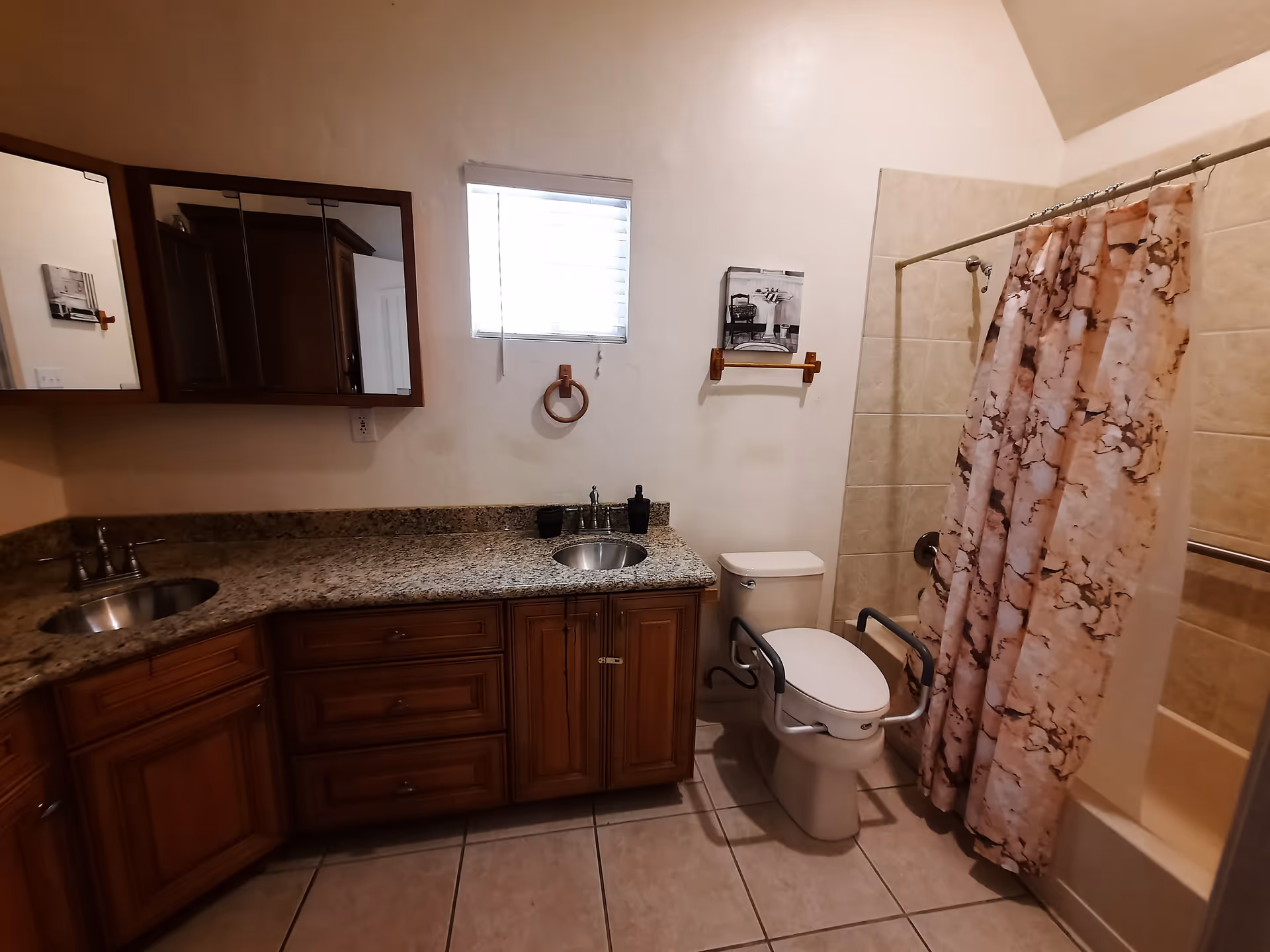 A bathroom with a granite countertop featuring two sinks and wooden cabinets underneath. There is a window with blinds above the countertop, a toilet with safety rails on both sides, and a bathtub with a pink and brown patterned shower curtain. The walls are light-colored, and the floor is tiled.