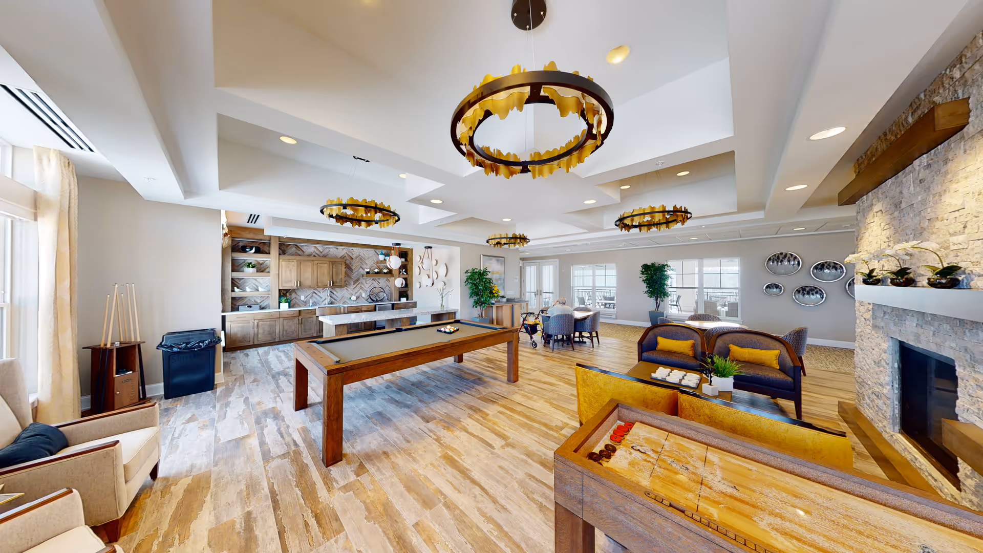 A spacious senior living community room featuring a pool table in the center, comfortable seating areas with sofas and armchairs, a stone fireplace on the right, and a kitchenette with wooden cabinets and a herringbone tile backsplash in the background. The room is well-lit with large windows and decorative circular chandeliers hanging from the ceiling.