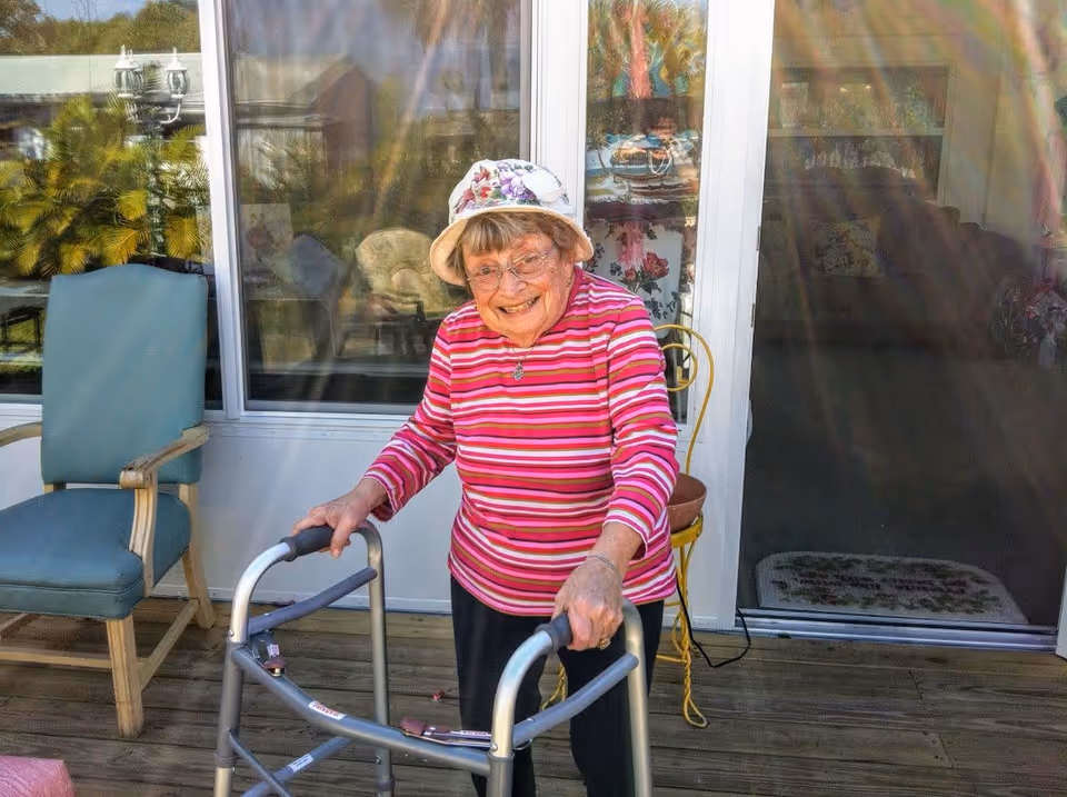 An elderly woman wearing a floral hat and a pink striped long-sleeve shirt is smiling while using a walker on a wooden deck outside a building with large glass doors and windows. There is a blue cushioned chair and a yellow metal chair nearby.
