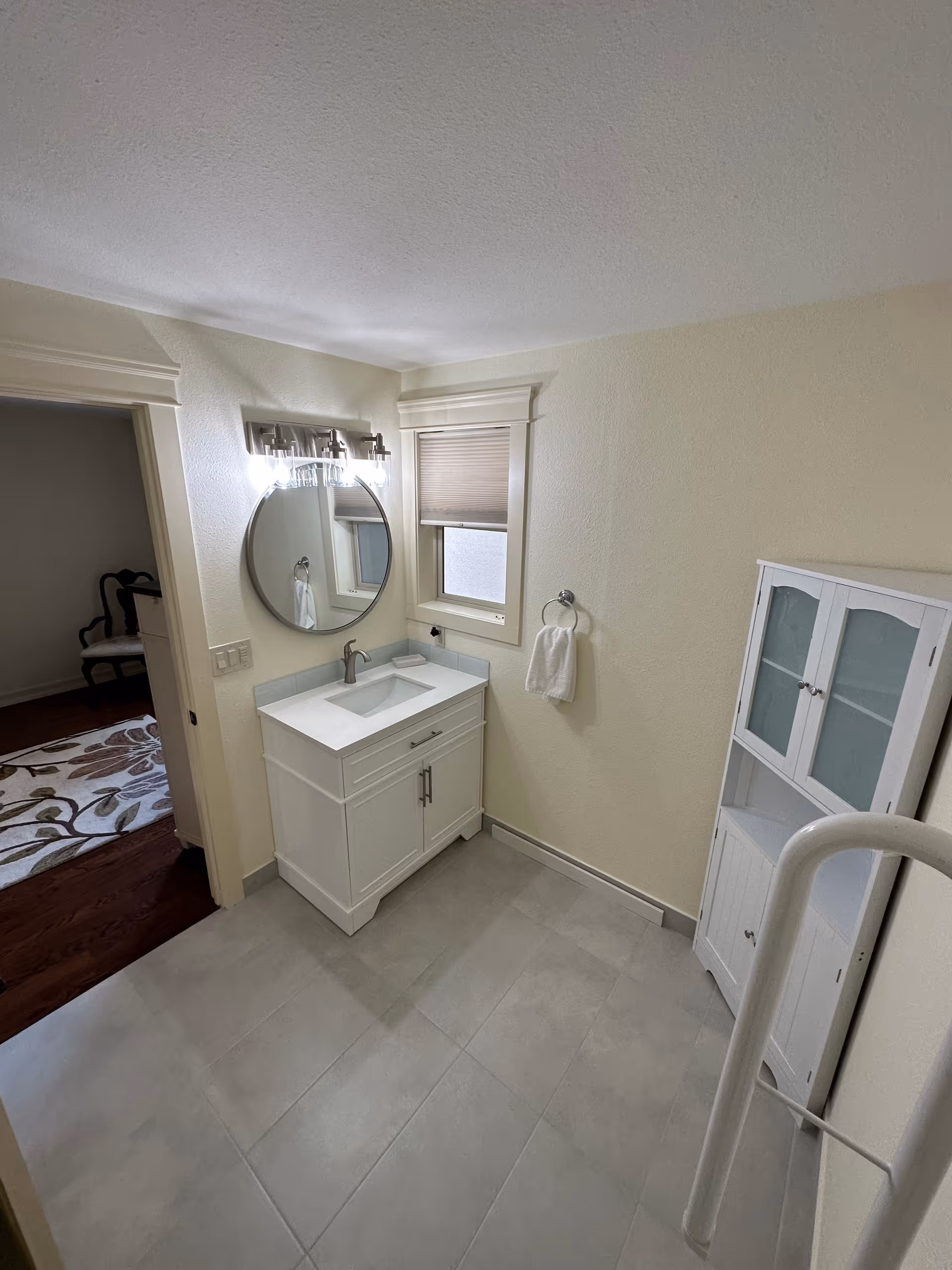 A clean bathroom with light-colored walls and floor tiles. There is a white vanity with a sink and a round mirror above it, illuminated by a light fixture. A small window with a blind is next to the mirror. A white towel hangs on a ring on the wall. A white cabinet with glass doors stands against the right wall. The bathroom opens to an adjacent room with a wooden floor and a patterned rug.