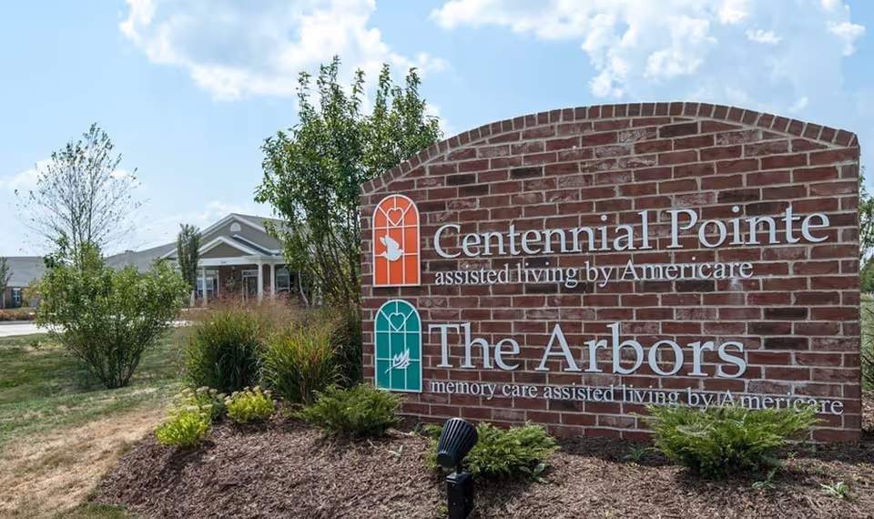 A brick sign for Centennial Pointe assisted living by Americare and The Arbors memory care assisted living by Americare, surrounded by landscaping with bushes and trees, with a building visible in the background under a partly cloudy sky.