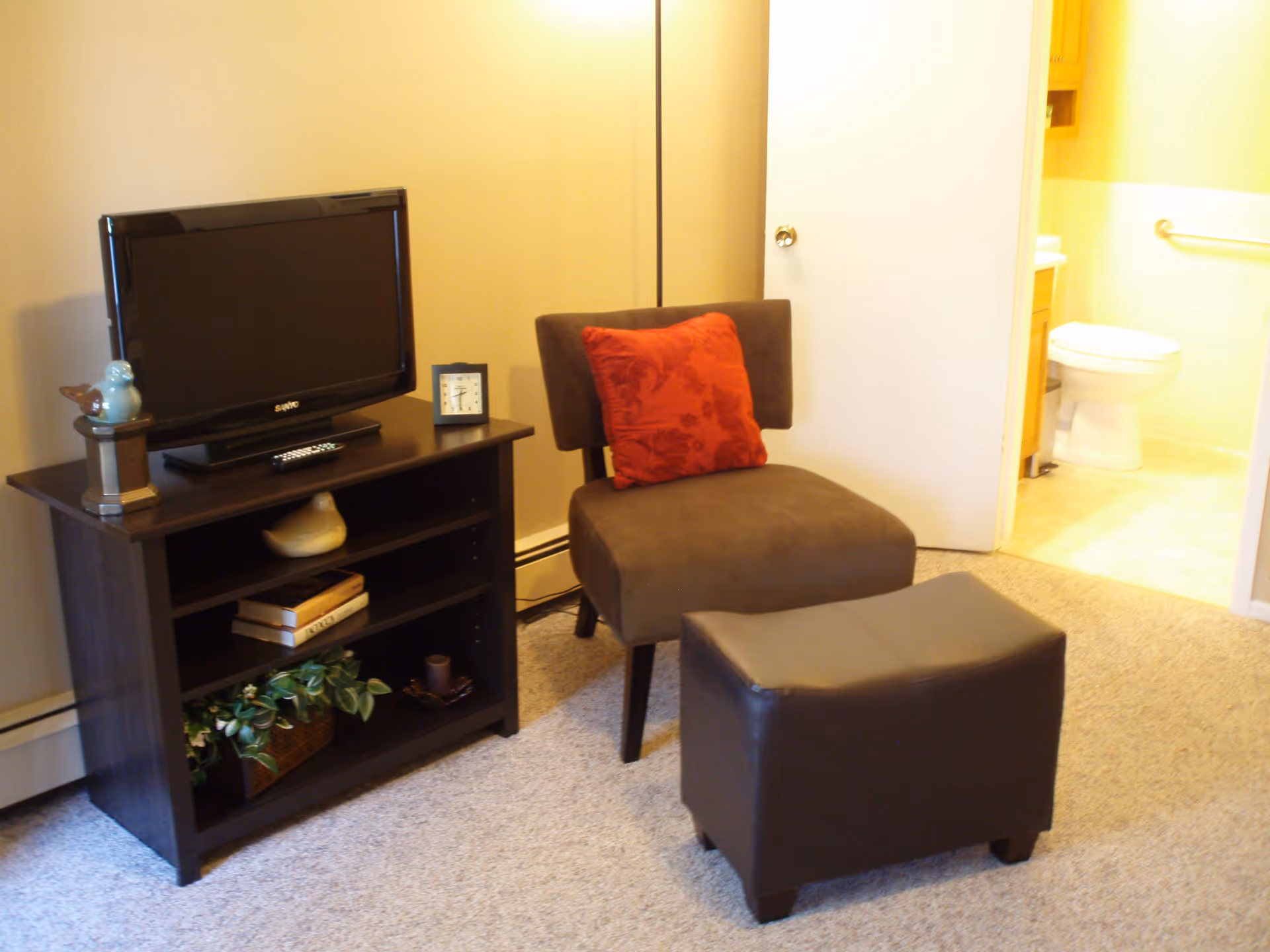 A small living area with a brown upholstered chair featuring a red cushion, a matching brown ottoman, and a dark wooden TV stand holding a flat-screen TV, a clock, decorative birds, books, and a plant. An open door reveals a bathroom with a toilet and sink.