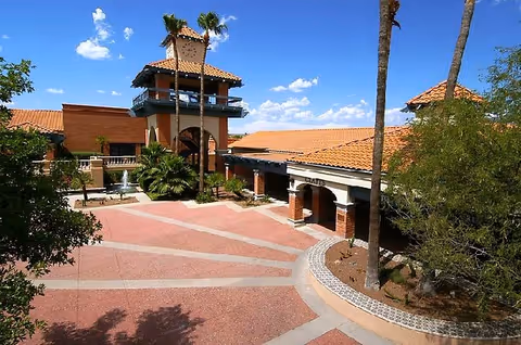 Exterior view of Sun City Oro Valley Welcome Center featuring a courtyard with a fountain, palm trees, and a building with terracotta roof tiles under a blue sky with scattered clouds.