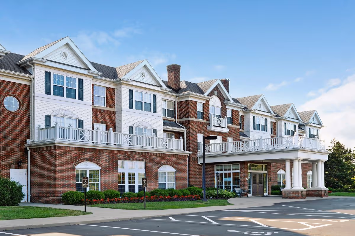 Exterior view of a multi-story senior living facility building with red brick and white siding, featuring multiple windows, balconies, and a covered entrance with white columns. The foreground shows a parking area with designated handicap parking spaces and landscaped greenery along the building.