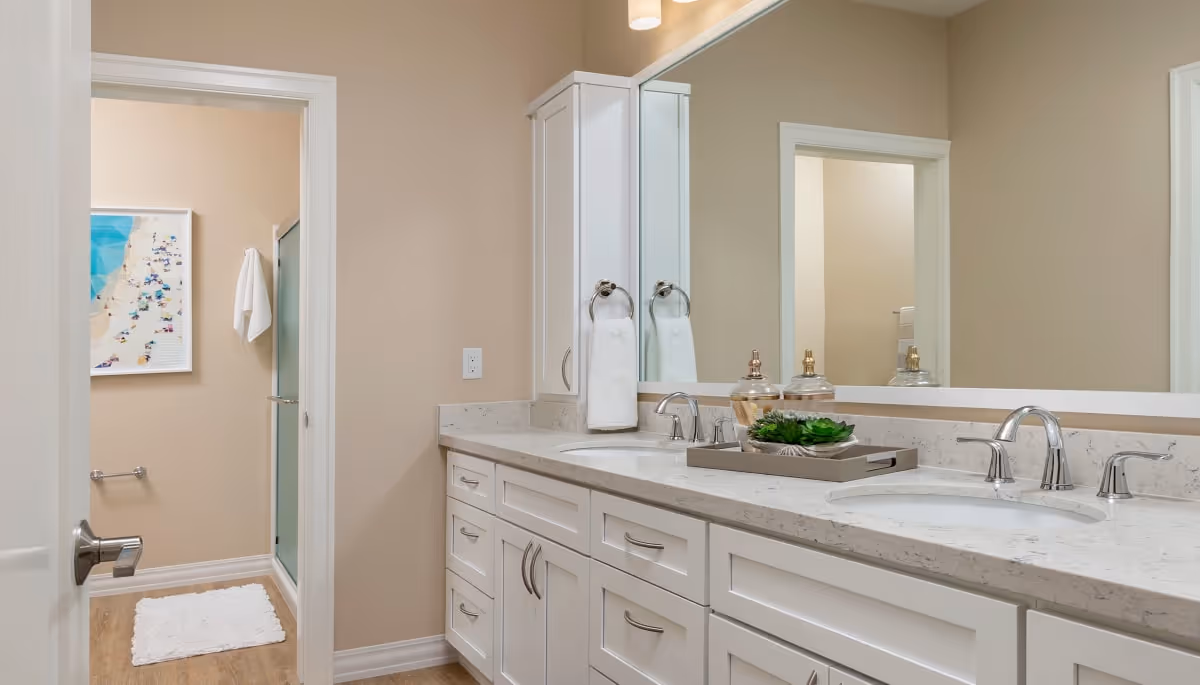 Bright bathroom with a double-sink vanity, large mirror, white cabinets, and a doorway leading to a shower.