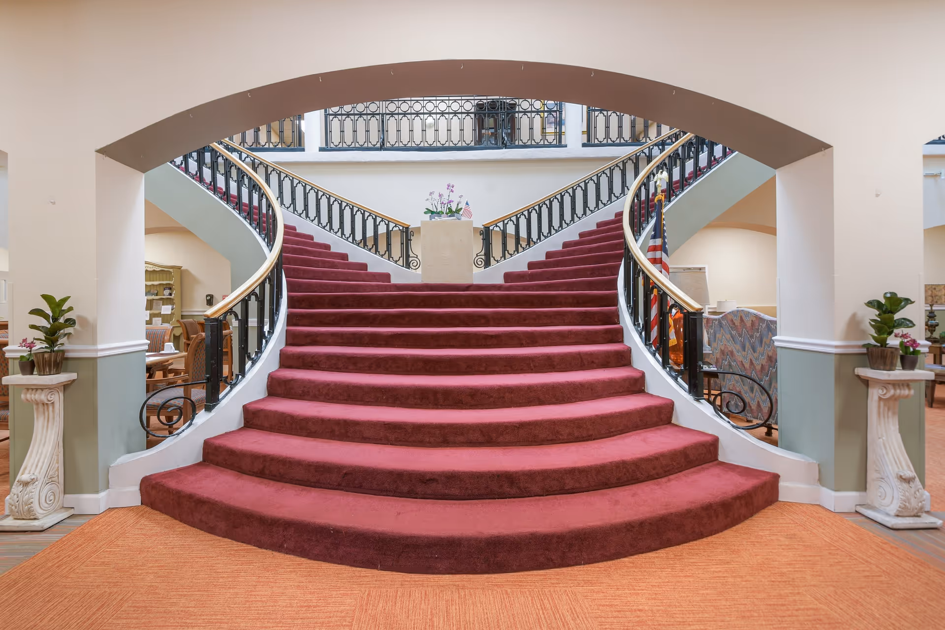 A grand, curved staircase with red carpeting and black railings with gold handrails in a senior living community. The staircase splits into two directions at the top, with a small pedestal holding a plant and an American flag in the center. The area around the staircase has seating areas with chairs and tables, and decorative plants on pedestals on either side of the staircase base.
