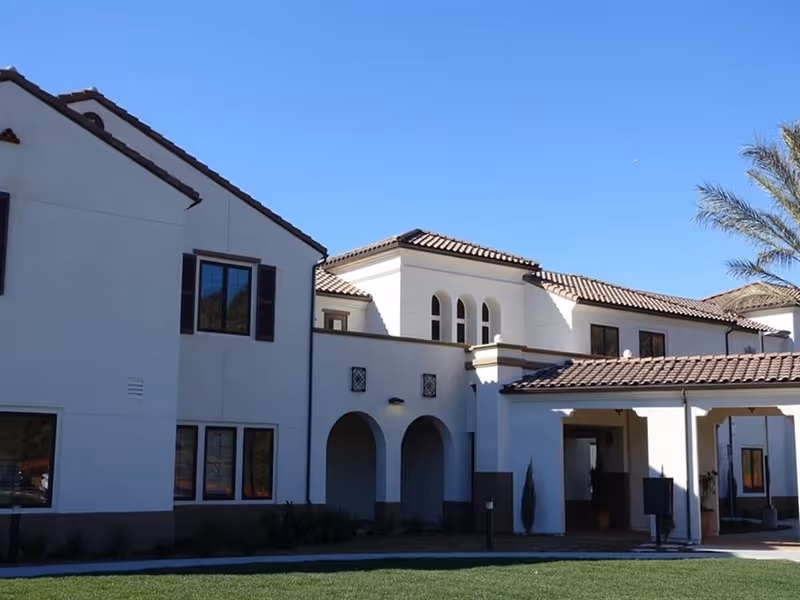 Exterior view of a two-story building with white walls and brown tiled roof under a clear blue sky, featuring arched doorways and several windows, with a green lawn in the foreground and a palm tree on the right side.