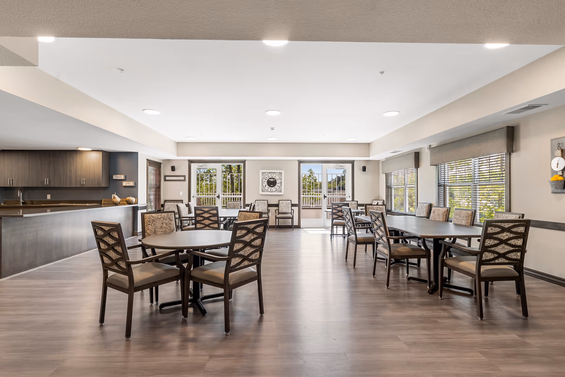 A spacious senior living dining room with multiple tables and chairs arranged neatly. The room features large windows with blinds allowing natural light to fill the space. There is a kitchen area with dark cabinetry and a countertop on the left side. The flooring is wood-style, and the walls are painted in neutral tones. The room has ceiling lights and a clock on the far wall.