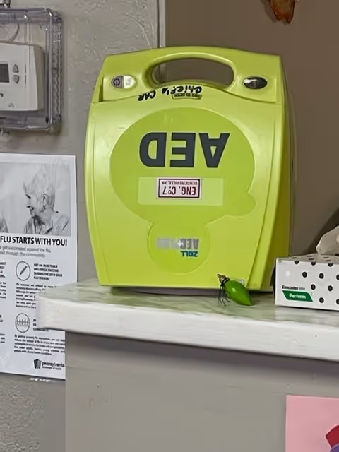 A bright yellow AED (Automated External Defibrillator) device mounted on a wall above a white counter. To the left of the AED, there is a health information poster about flu prevention and a thermostat on the wall. On the counter below the AED, there is a small green decorative item and a white box with black dots labeled 'Cascade Pureform'.