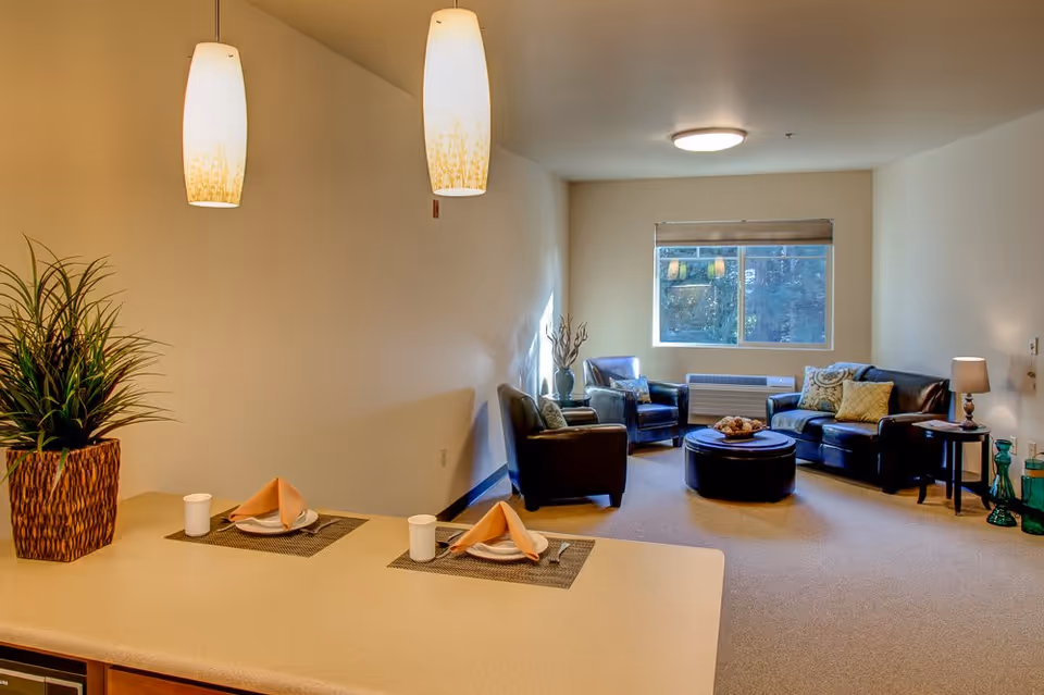 A cozy living room and dining area in an assisted living facility. The living room features a black leather sofa, two black armchairs, a round ottoman, and a window with a view outside. The dining area has a countertop with two place settings, each with a plate, napkin, and cup. Two pendant lights hang above the countertop, and a potted plant is placed on the counter.