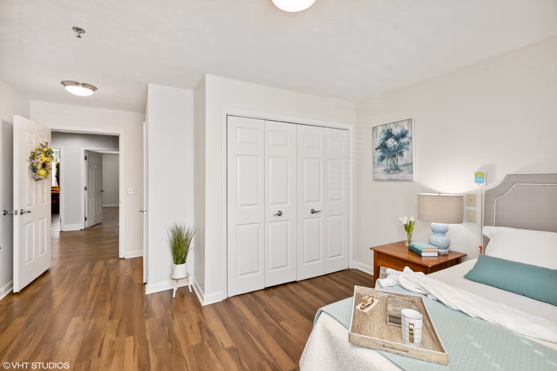 A bright and clean bedroom with a bed featuring a light gray upholstered headboard, white and teal bedding, and a wooden tray with a cup and glasses on it. Next to the bed is a wooden nightstand with a blue lamp, books, and a small vase with white flowers. The room has wood flooring, white walls, a closet with double doors, a small potted plant on a stand, and an open door leading to a hallway.