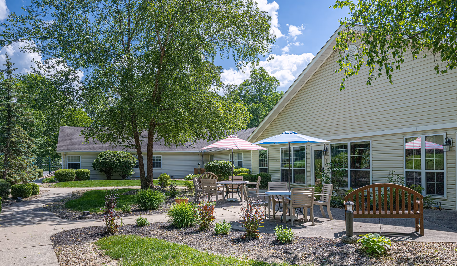 Outdoor patio area at Arden Courts - ProMedica Memory Care Community (Parma) featuring several tables with umbrellas, chairs, a wooden bench, and landscaped garden beds with shrubs and small plants. The patio is adjacent to a light-colored building with large windows, surrounded by trees and greenery under a partly cloudy blue sky.
