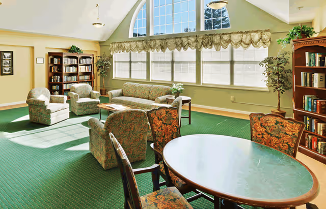 A bright and spacious common area with green carpet, floral upholstered chairs, a matching sofa, and wooden bookshelves filled with books. Large windows with white blinds and a decorative valance let in natural light. A round table with floral upholstered chairs is in the foreground, and potted plants add greenery to the room.