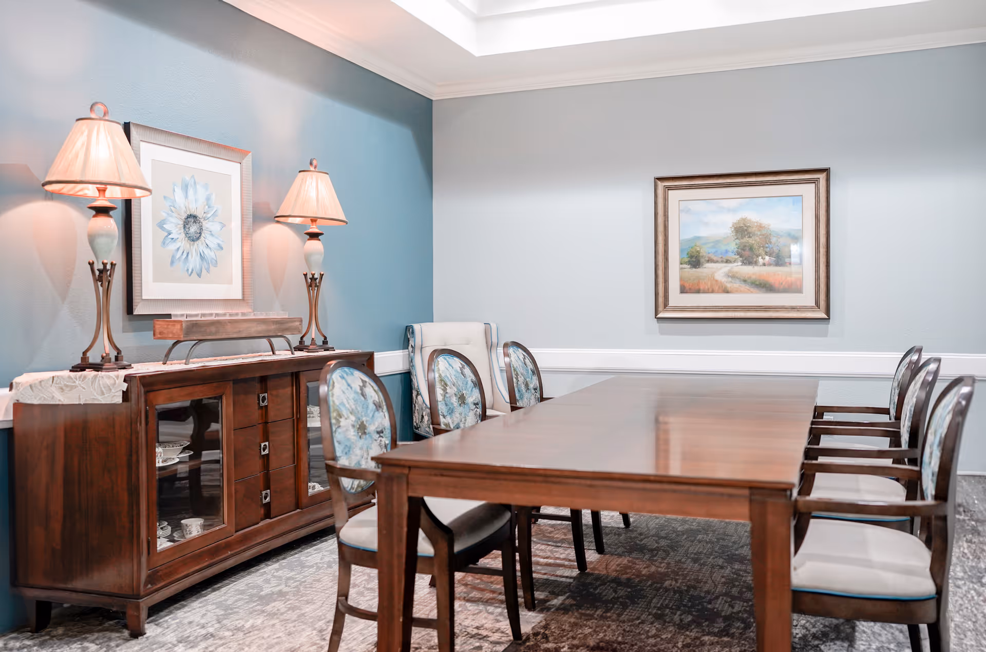 A dining room with a long wooden table surrounded by six chairs with floral upholstery. Against the wall is a wooden sideboard with glass doors displaying dishes, topped with two table lamps and a framed flower artwork. Another framed landscape painting hangs on the opposite wall. The walls are painted in light blue and gray tones.