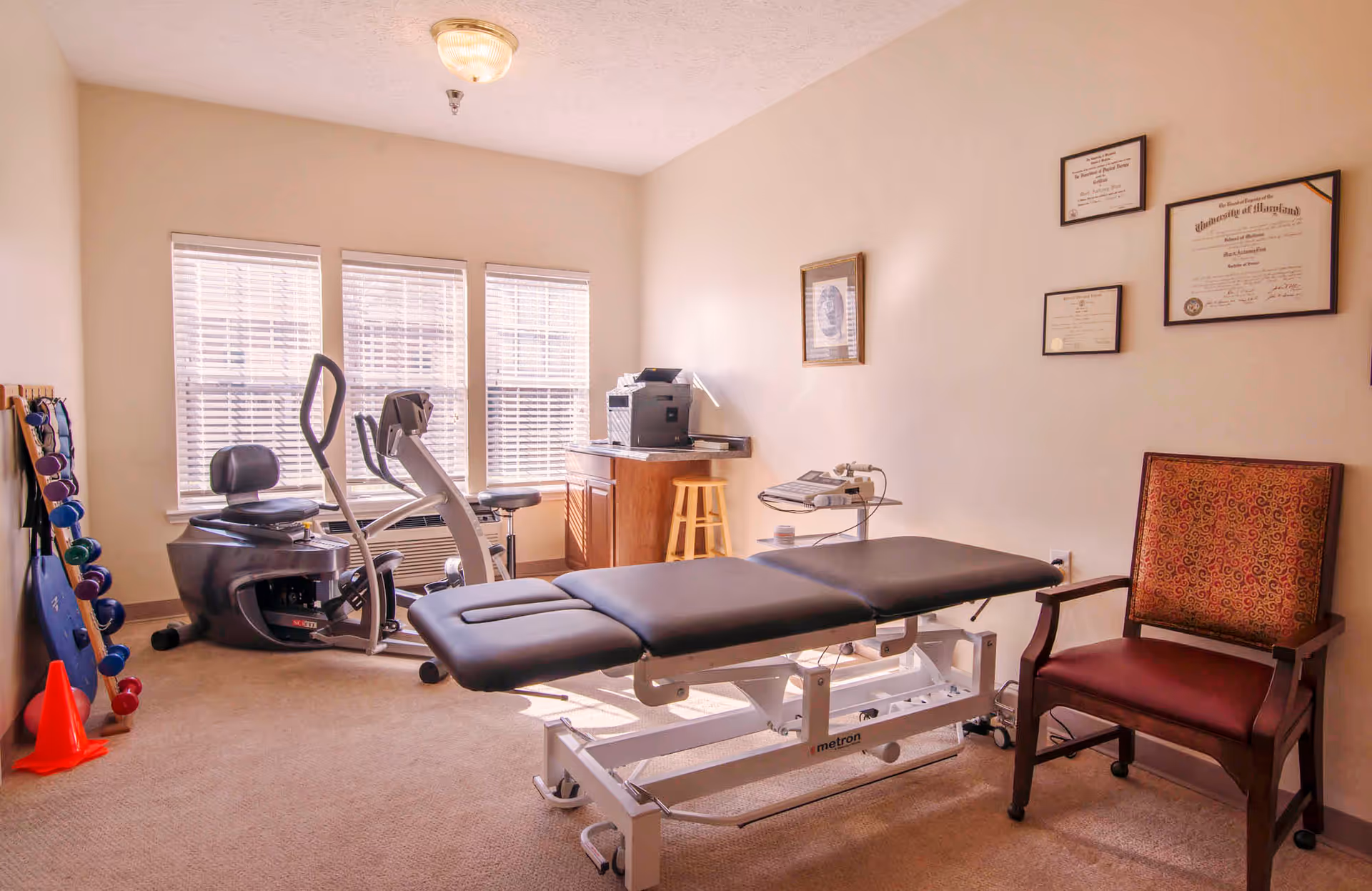 A therapy or rehabilitation room with a black padded treatment table in the center, exercise equipment including a recumbent bike and elliptical near three windows with blinds, a rack of colorful dumbbells and exercise cones on the left, a wooden cabinet with a printer and stool, a red cushioned chair, and framed certificates on the wall.