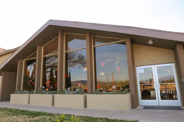 Exterior view of a building with large vertical windows decorated with heart-shaped stickers. The building has a slanted roof and double glass doors. There are planters with greenery in front of the windows and a grassy area in the foreground.