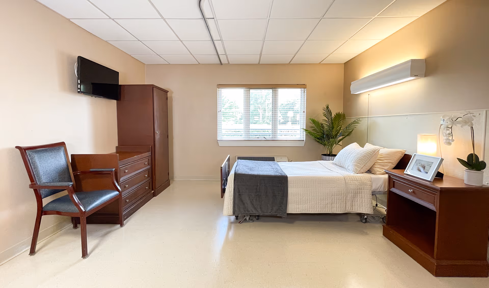 A clean nursing facility patient bedroom with a single bed, nightstand, dresser, chair, wall-mounted TV and a window.