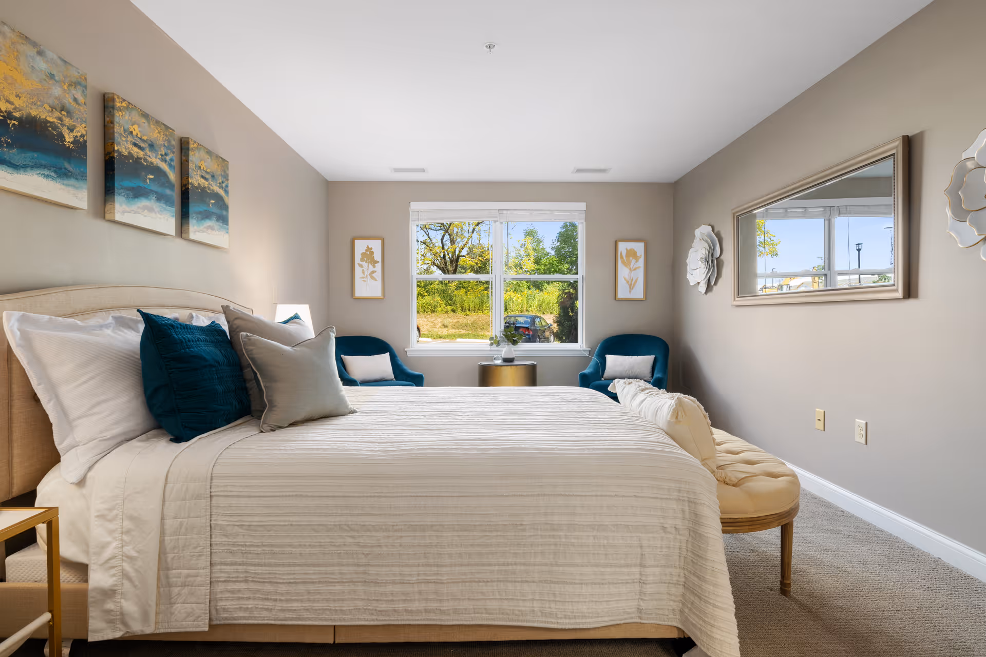 Sunlit bedroom featuring a neatly made bed with teal accent pillows, two teal chairs by a window, and neutral wall decor.