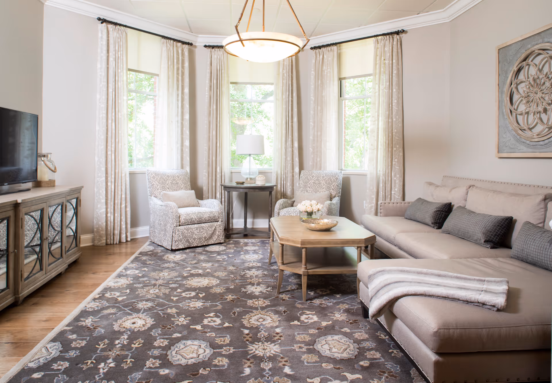 A bright and cozy living room with a large beige sectional sofa adorned with gray cushions and a striped throw blanket. Two patterned armchairs are positioned near three tall windows with light curtains. A wooden coffee table with decorative items sits on a patterned area rug. A wooden TV stand with a flat-screen TV is on the left side, and a round side table with a lamp is between the armchairs. A decorative wall art piece hangs above the sofa.