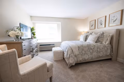 A cozy bedroom with a large bed covered in patterned bedding and multiple pillows. There is a cushioned ottoman at the foot of the bed, a wooden dresser with a flat-screen TV on top, and a comfortable armchair. The walls are light-colored with three framed botanical prints hanging above the bed. A window allows natural light to brighten the room.