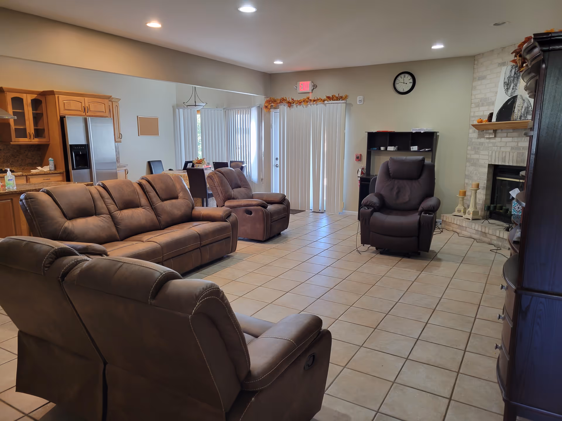 A spacious senior living common area with brown leather recliner sofas and chairs arranged on a tiled floor. The room features a brick fireplace with decorative candles and artwork above it. In the background, there is a kitchen area with wooden cabinets and a stainless steel refrigerator, as well as a dining table with chairs near vertical blinds covering glass doors. The walls are light-colored, and there is a clock and an exit sign visible.