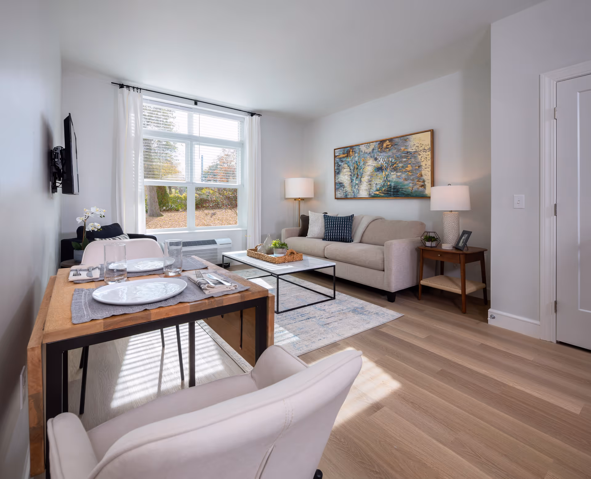 A bright and modern living room with light wood flooring, a beige sofa with cushions, a wooden side table with a lamp and picture frame, a coffee table with decorative items, a wall-mounted TV, and a small dining table set for two near a large window with white curtains.