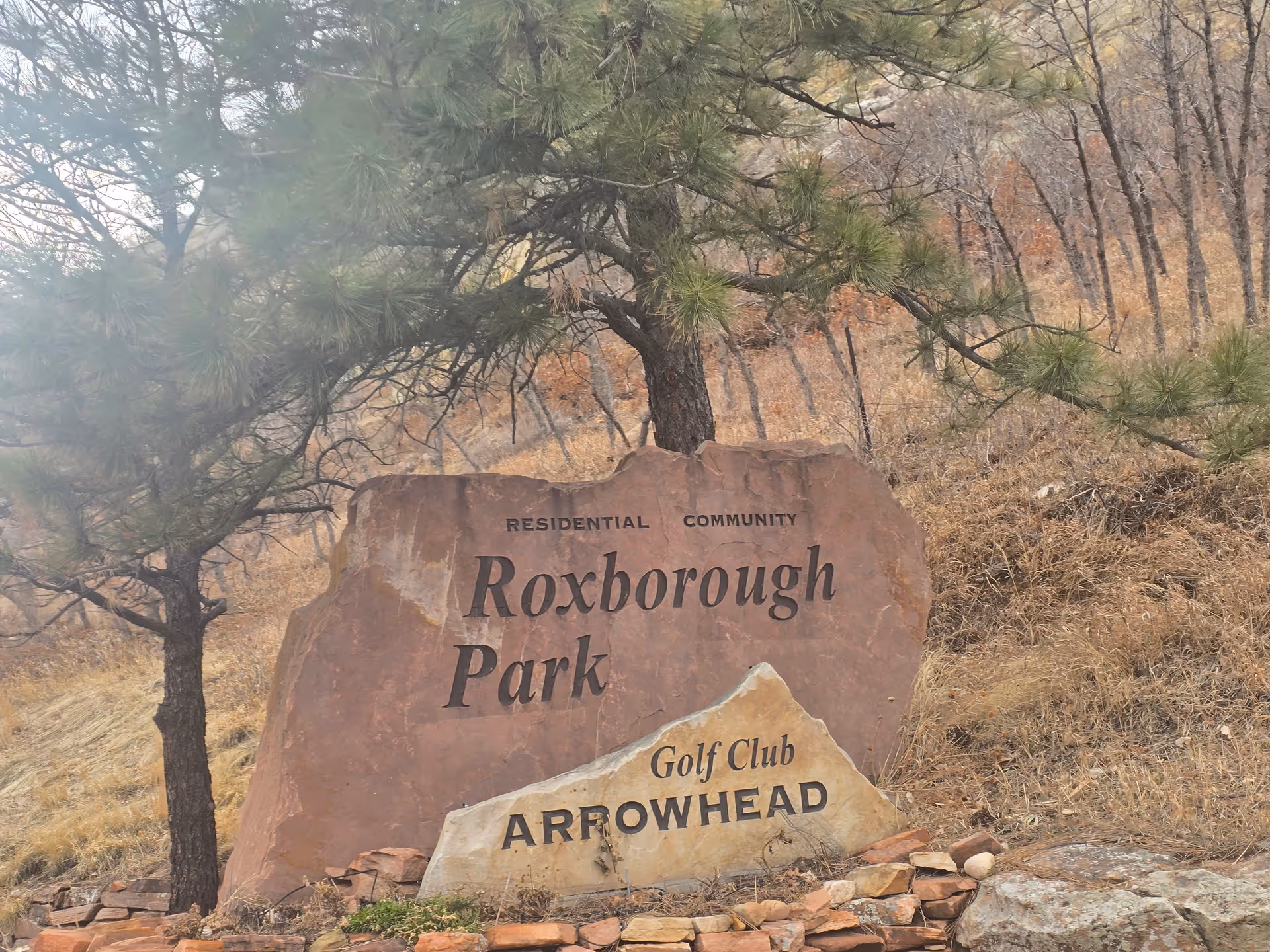 Large stone sign in a natural outdoor setting with trees and dry grass. The sign reads 'Residential Community Roxborough Park' and a smaller stone in front reads 'Golf Club Arrowhead'.