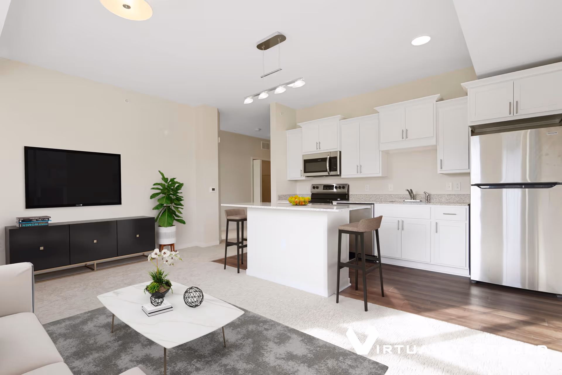 A modern open-concept living area and kitchen with white cabinetry, stainless steel appliances, a kitchen island with two bar stools, a wall-mounted flat-screen TV above a black media console, a small indoor plant, and a coffee table with decorative items on a gray rug.