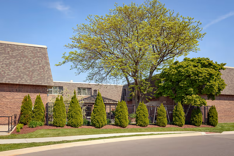 Exterior view of a senior living facility with a row of neatly trimmed evergreen shrubs and two large leafy trees in front of a brick building under a clear blue sky.