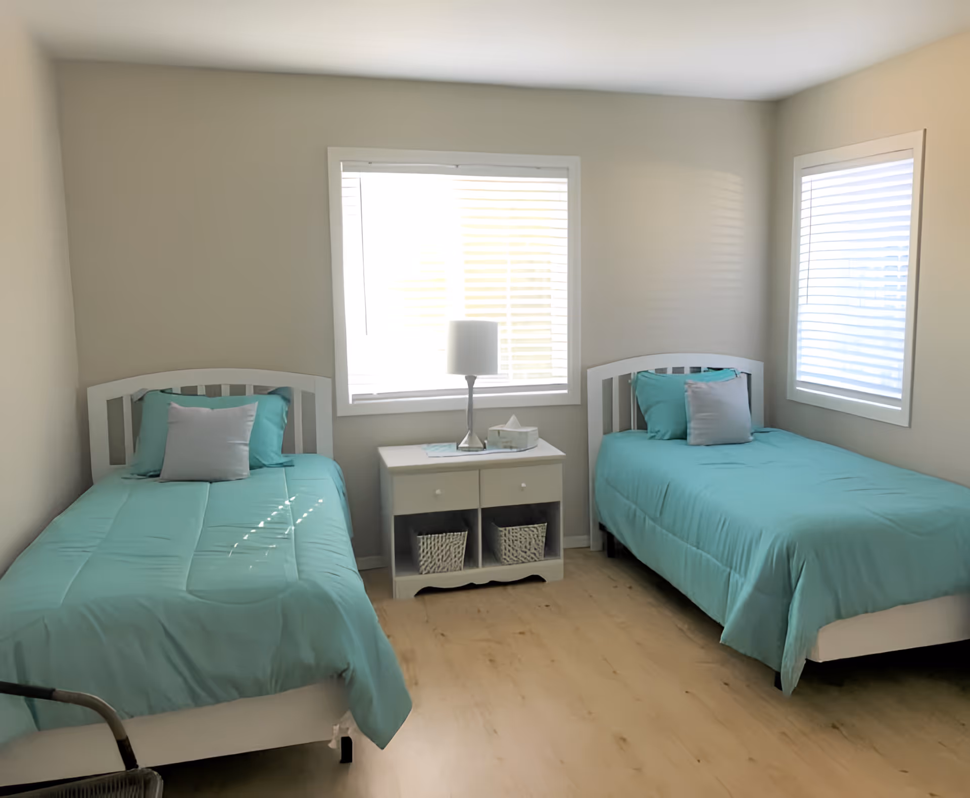 A simple bedroom with two single beds, each with teal bedding and a gray pillow. Between the beds is a white nightstand with two drawers and two baskets underneath. On top of the nightstand is a lamp and a tissue box. The room has light-colored walls and two windows with white blinds, allowing natural light to enter.