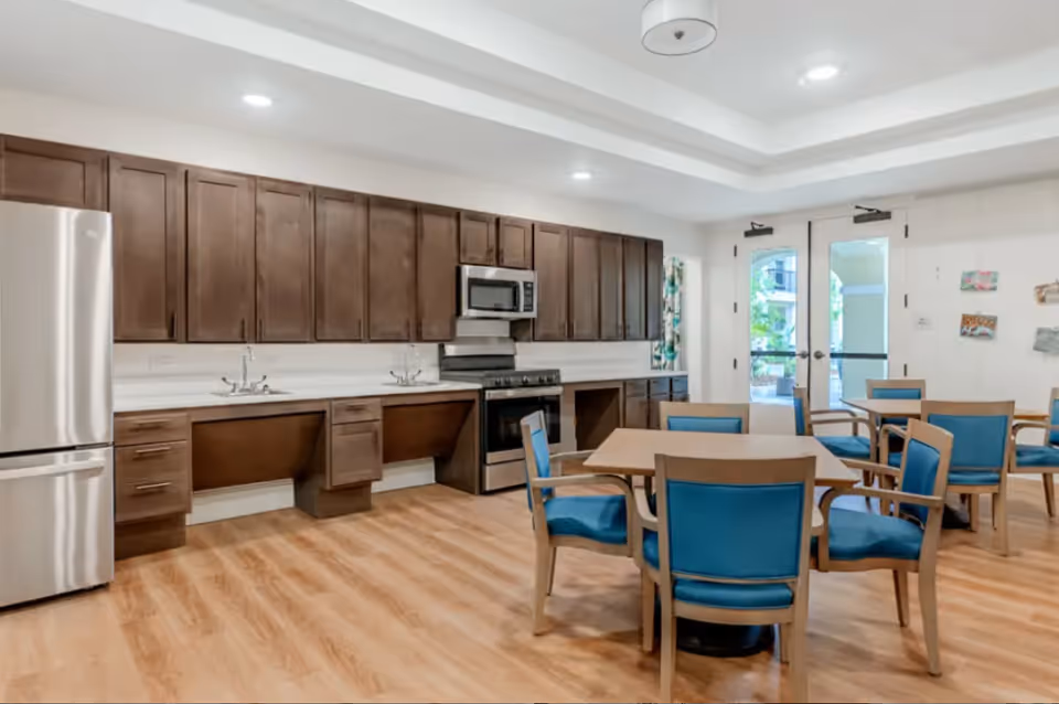 A spacious kitchen and dining area with wooden cabinets, stainless steel refrigerator, stove, and microwave. The room has light wood flooring, multiple tables with blue cushioned chairs, and double glass doors leading outside.