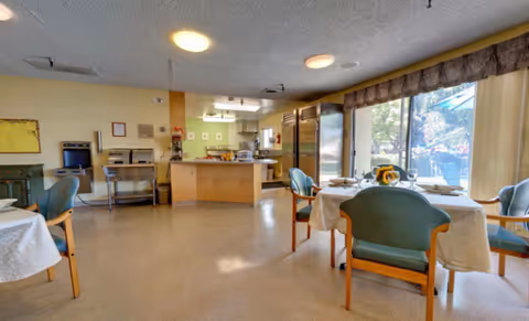 A bright dining area in a senior living facility with tables covered in white tablecloths, set with plates and utensils. There are green cushioned chairs around the tables. The room has large windows with vertical blinds letting in natural light and a view of an outdoor garden. In the background, there is a kitchen area with stainless steel appliances and a countertop.
