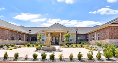Front exterior view of a single-story brick building with a covered entrance, two lampposts, landscaped garden with small bushes, and a clear blue sky with some clouds.