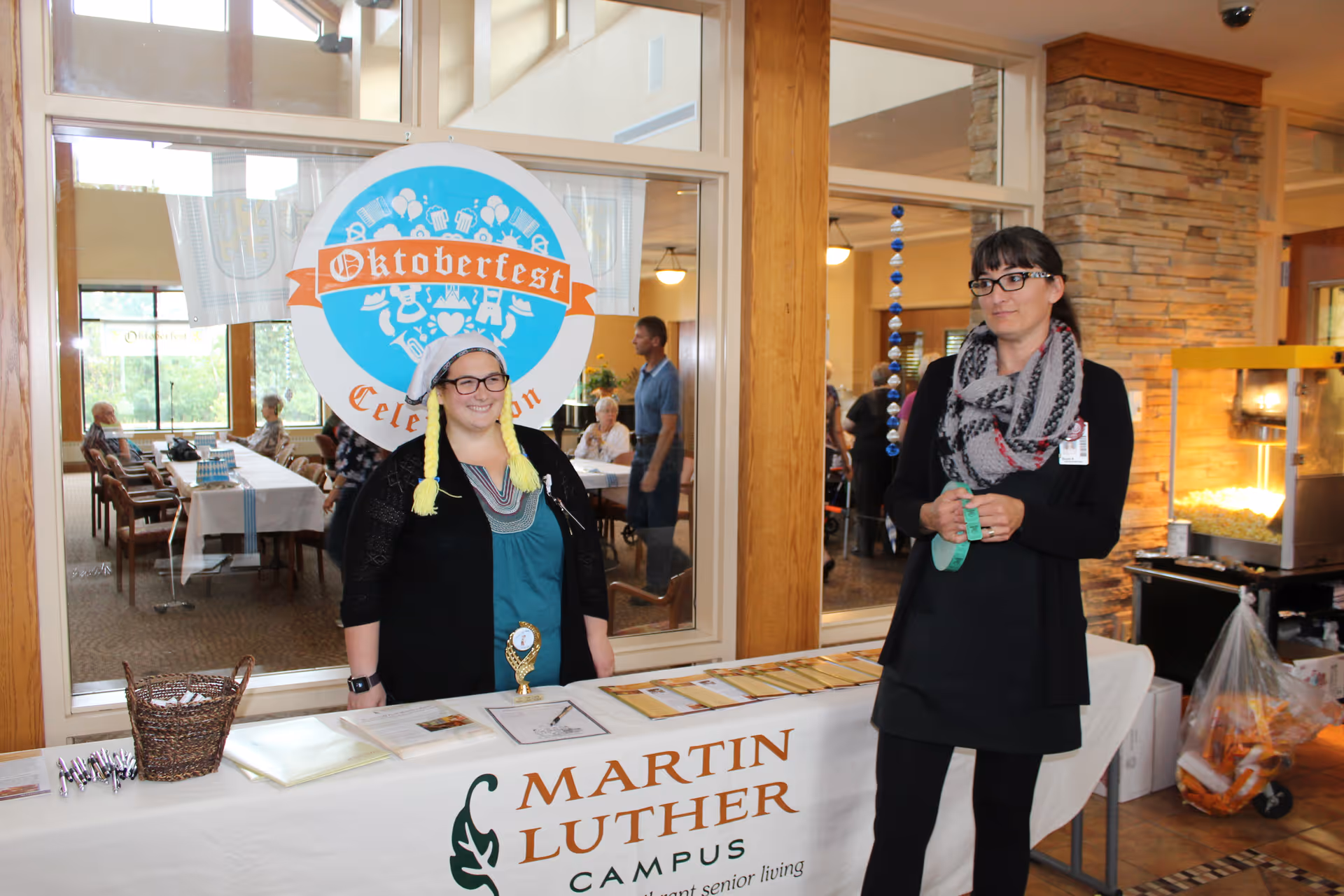 Two women standing behind a table covered with a white cloth that has the Martin Luther Campus logo and name. One woman is wearing a hat with yellow braids and glasses, smiling, while the other woman is wearing glasses and a scarf, holding green tickets. Behind them is a glass window showing a dining area with elderly people seated at tables. There is a large Oktoberfest Celebration sign on the window and a popcorn machine to the right.