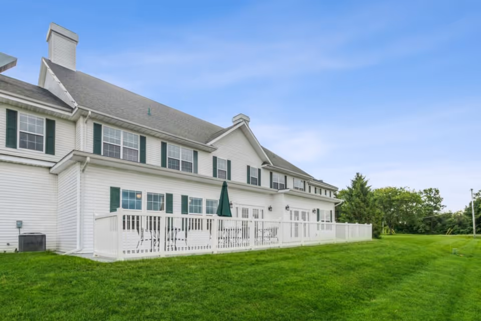 White two-story building with green shutters, a fenced patio and a wide grassy lawn under a blue sky.