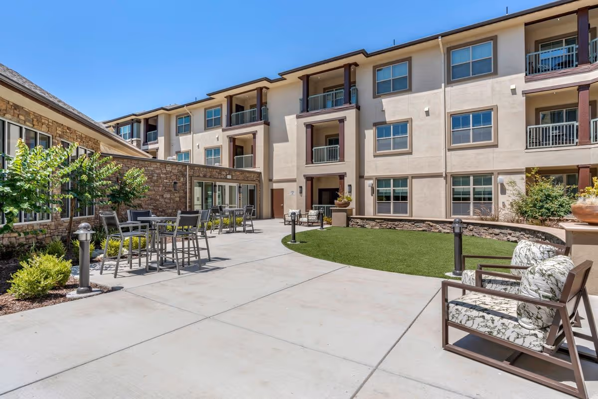 Outdoor courtyard of a multi-story senior living building with seating, tables, and a small lawn.