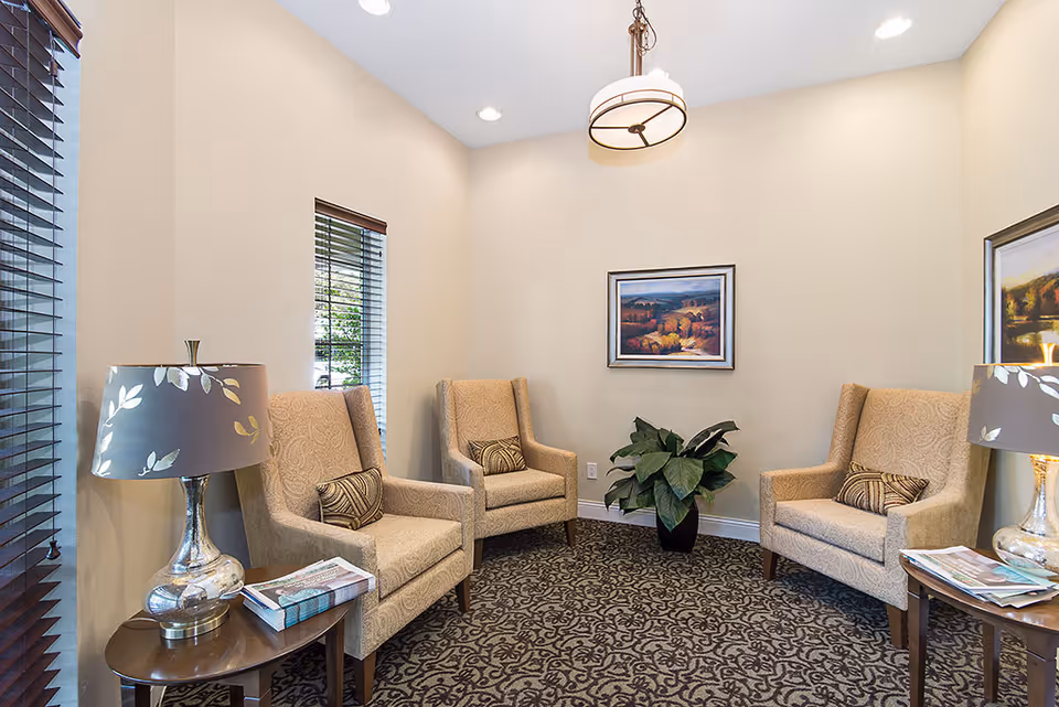 A cozy sitting area with three beige upholstered armchairs arranged in a semi-circle on a patterned carpet. Each chair has a decorative pillow. There are two small wooden side tables with silver lamps featuring leaf-patterned shades. A potted plant is placed against the wall beneath a framed landscape painting. The room has beige walls, two windows with dark blinds, and a ceiling light fixture.