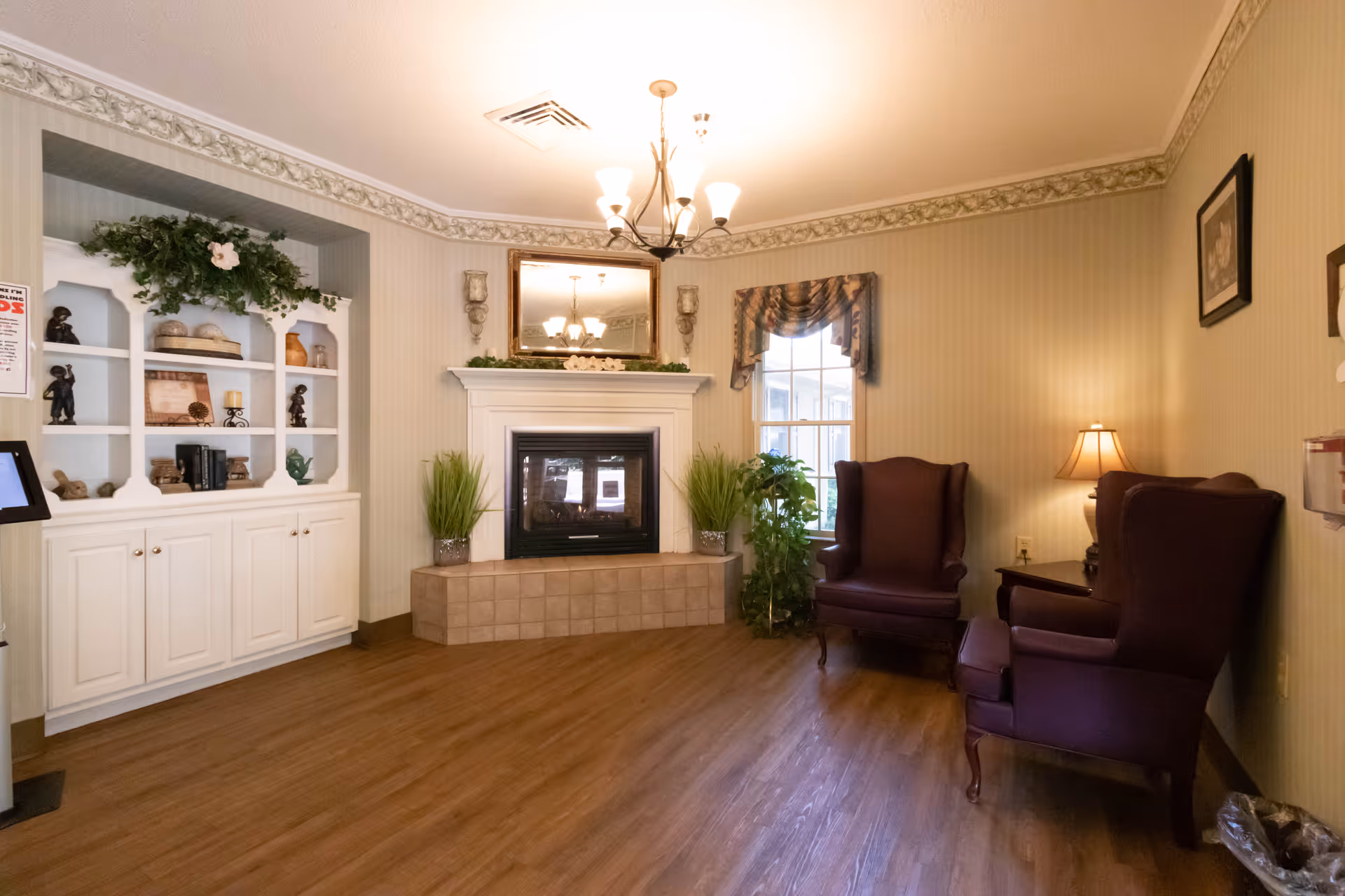 A cozy living room area with two dark purple wingback chairs, a small side table with a lamp, a window with a valance, a fireplace with a mirror above it, and built-in white shelves decorated with various items and greenery. The room has wood flooring and soft beige wallpaper with a floral border near the ceiling.