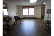 A spacious room with tiled flooring, two windows with blinds, and minimal furniture including two chairs, two small tables, a lamp, and a flower arrangement on one table.