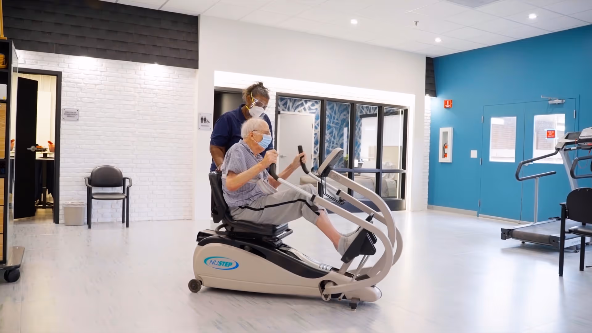 An elderly man wearing a mask is using a NuStep exercise machine in a bright rehabilitation room while a healthcare worker, also wearing a mask, stands behind him assisting. The room has white brick walls, a blue accent wall, and exercise equipment including a treadmill.