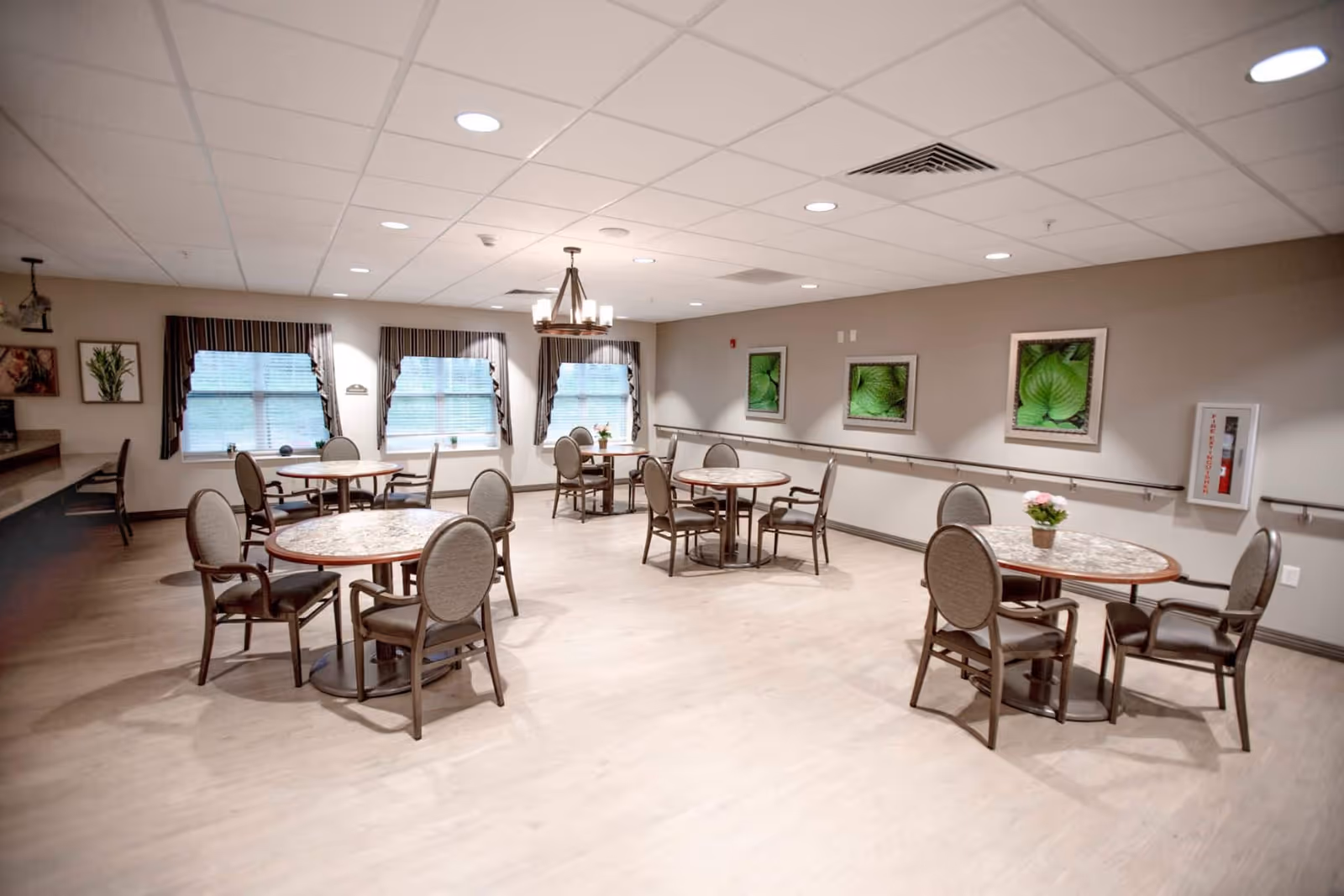 A bright and spacious dining room with several round tables, each surrounded by four chairs. The room has large windows with striped curtains, framed green leaf artwork on the walls, and a chandelier hanging from the ceiling. There is a fire extinguisher mounted on the wall and a counter with chairs along one side.