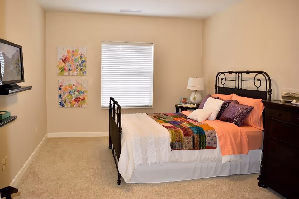 Furnished bedroom with an iron bed dressed in a colorful quilt, nightstand with lamp, dresser, wall-mounted TV, and a window with blinds.